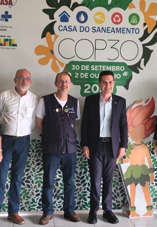 Três homens posam sorrindo diante de painel da Casa do Saneamento COP30 com ilustrações e logos coloridos.