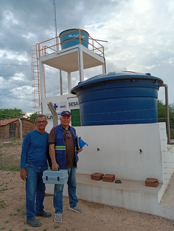Dois técnicos posam à frente de sistema de filtragem de água com grande reservatório azul e torre de suporte ao fundo.