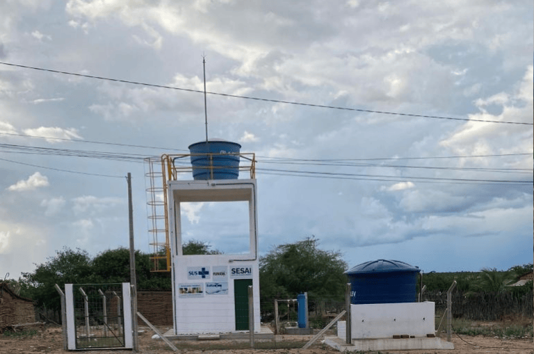 Unidade Salta-Z com torre branca, caixas d'água azuis e logos do SUS e SESAI sob céu nublado na aldeia Tuxi.