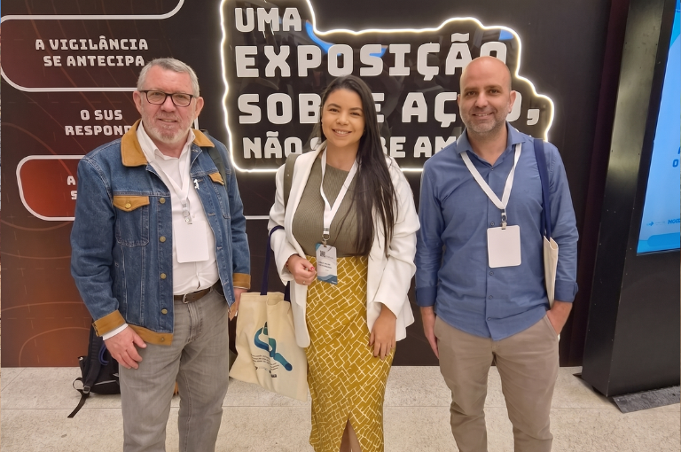 Três representantes da Funasa sorriem em pé na 18ª ExpoEpi, à frente de um painel preto com letreiro luminoso sobre vigilância e o SUS.