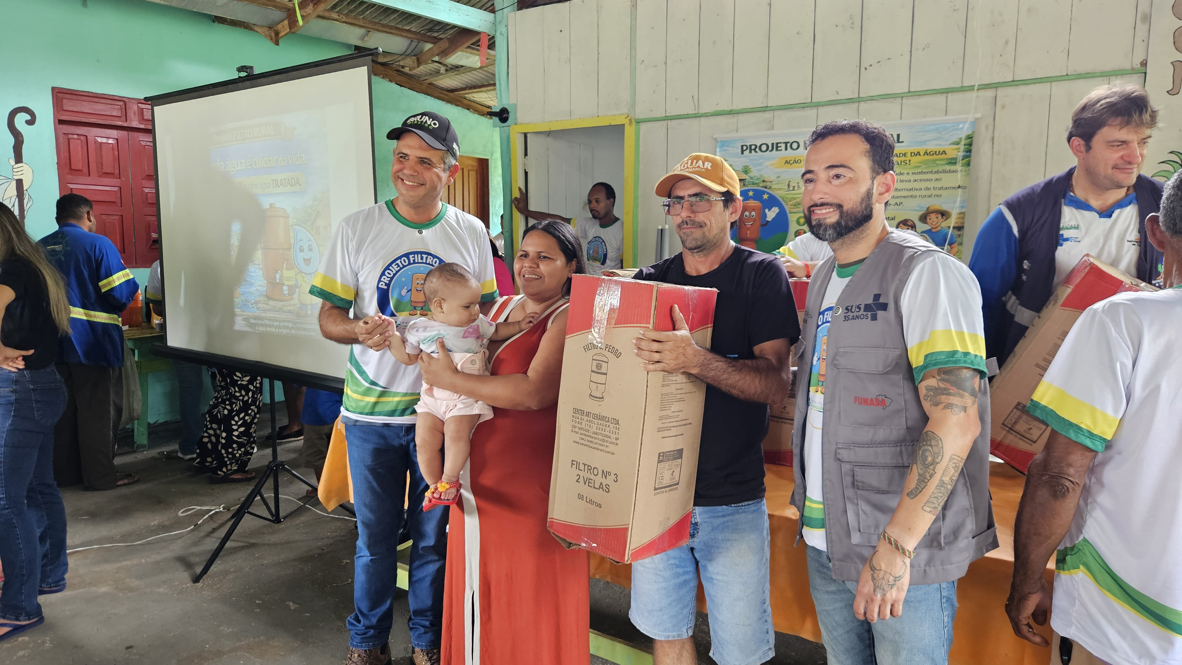Homens entregam um filtro de barro na caixa para uma mulher segurando um bebê em evento.