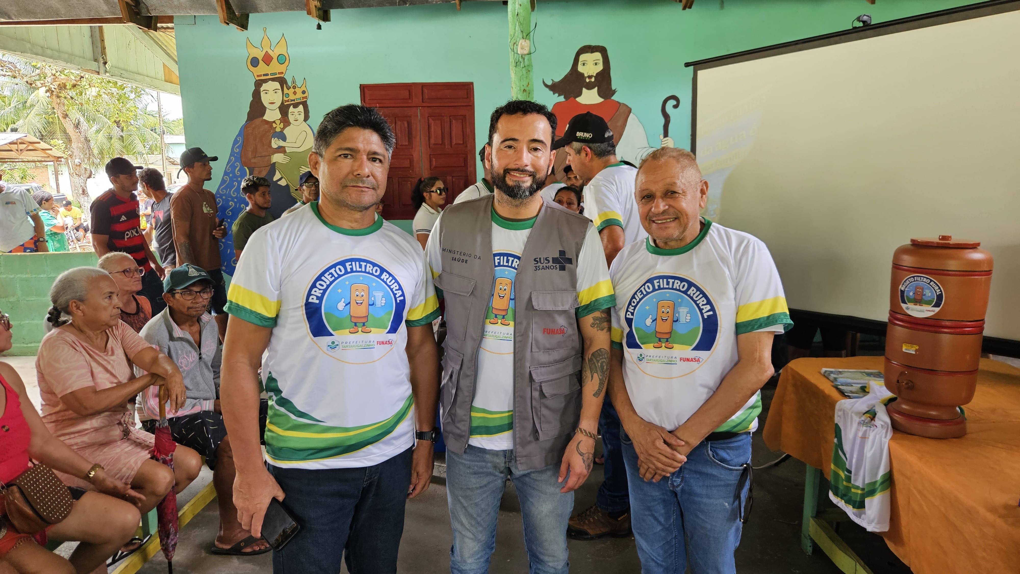 Três homens posam sorrindo na frente de um filtro de barro e pinturas religiosas na parede.