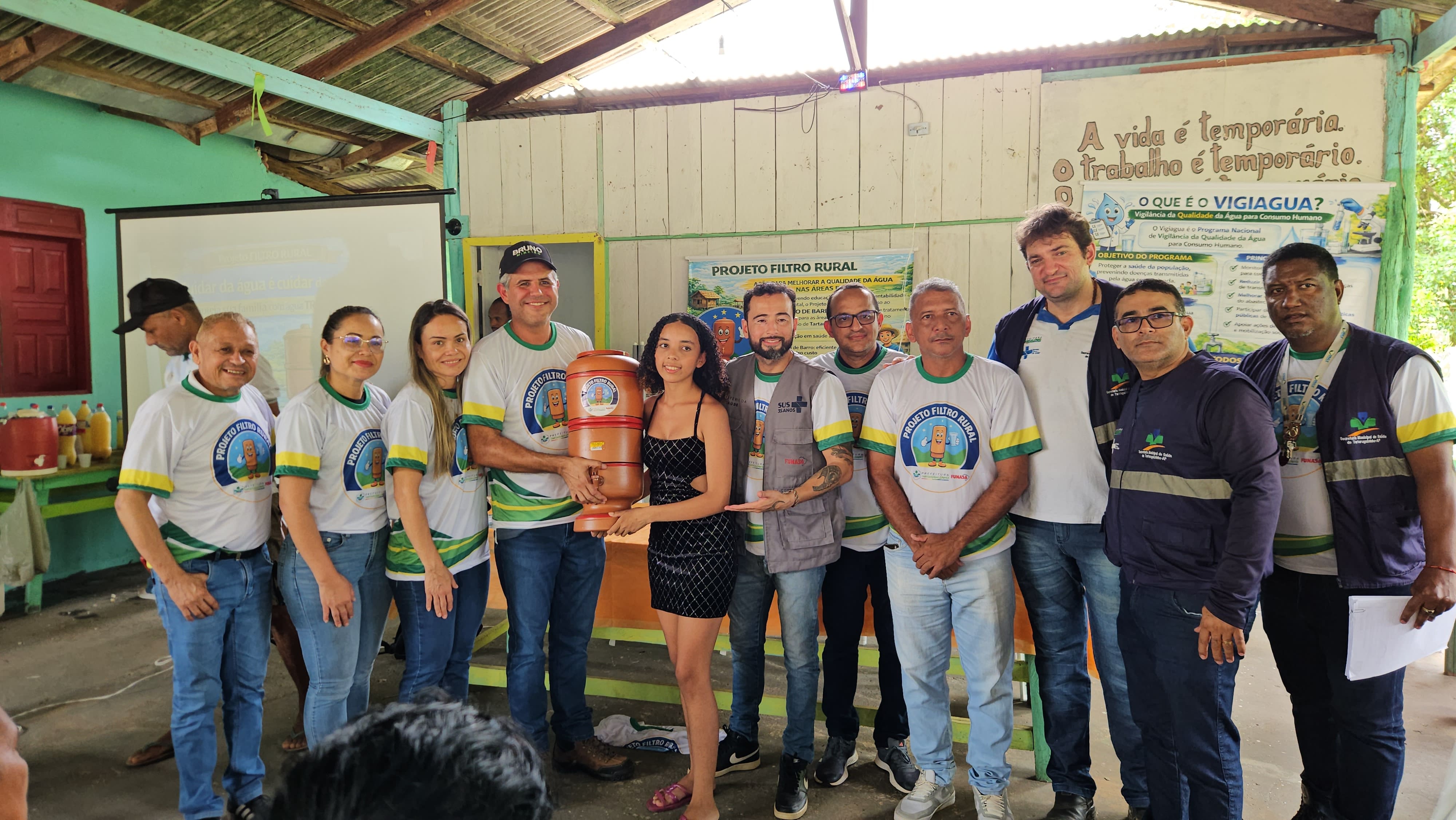 Grupo de pessoas posa com uma jovem que segura um filtro de barro em evento comunitário.