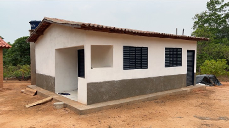 Lateral de casa branca recém-construída com telhado de cerâmica, janelas e portas de metal preto, sobre terreno de terra vermelha.