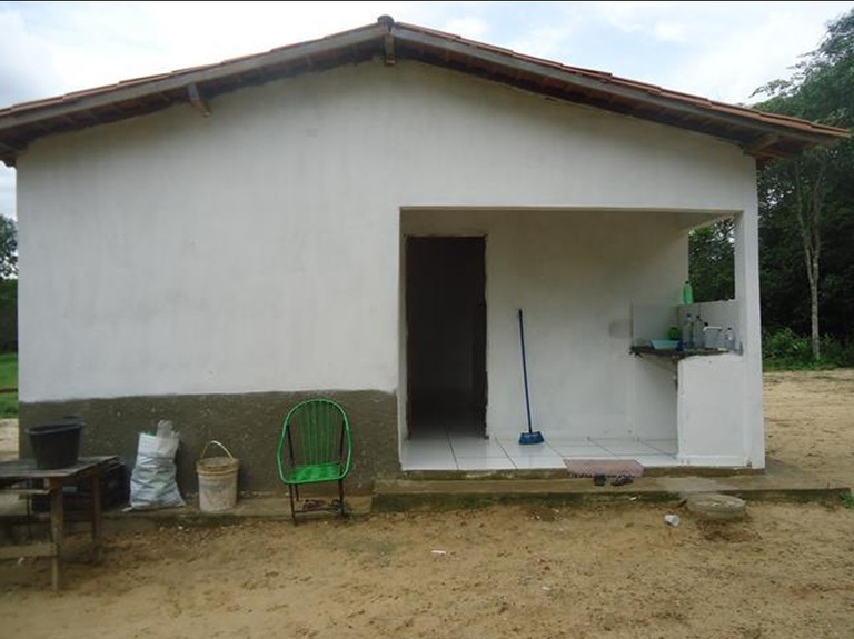 Varanda de casa simples com paredes brancas e base cinza, chão de cimento, uma cadeira verde de metal, uma vassoura e uma pia externa.