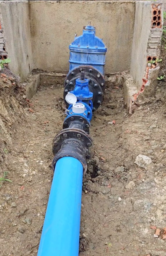 Tubulação azul conectada a válvulas metálicas pretas e registros de controle dentro de uma vala de concreto em canteiro de obras.