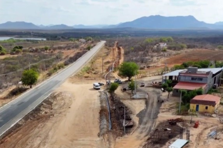 Vista aérea de estrada rural com vala de escavação lateral para instalação de adutora, cercada por vegetação baixa e montanhas ao fundo.