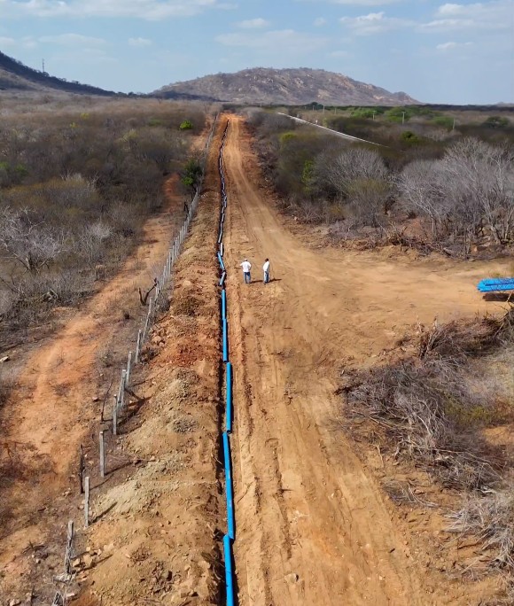 Vista aérea de extensa vala retilínea em estrada de terra com tubos azuis perfilados para instalação, cruzando área de vegetação seca.