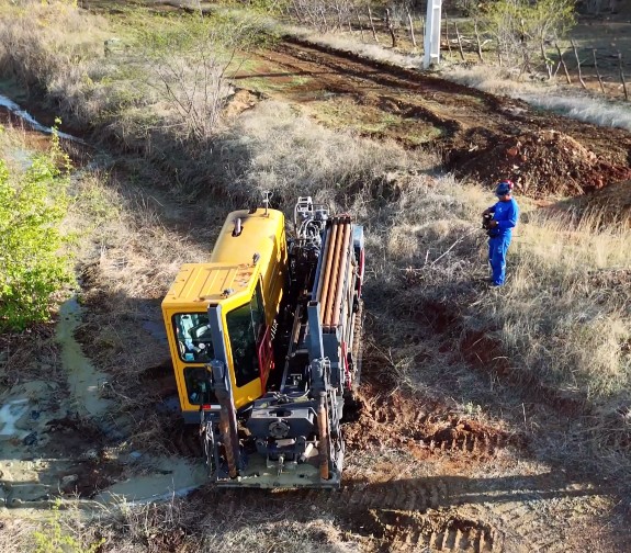 Vista aérea de máquina perfuratriz amarela operando em terreno de terra, com operário em uniforme azul ao lado monitorando a atividade.