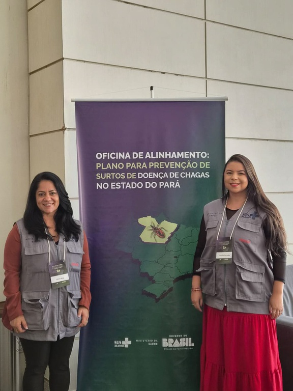 Duas mulheres sorriem ao lado de banner sobre o plano de prevenção da doença de Chagas. Ambas vestem coletes cinzas da Funasa.