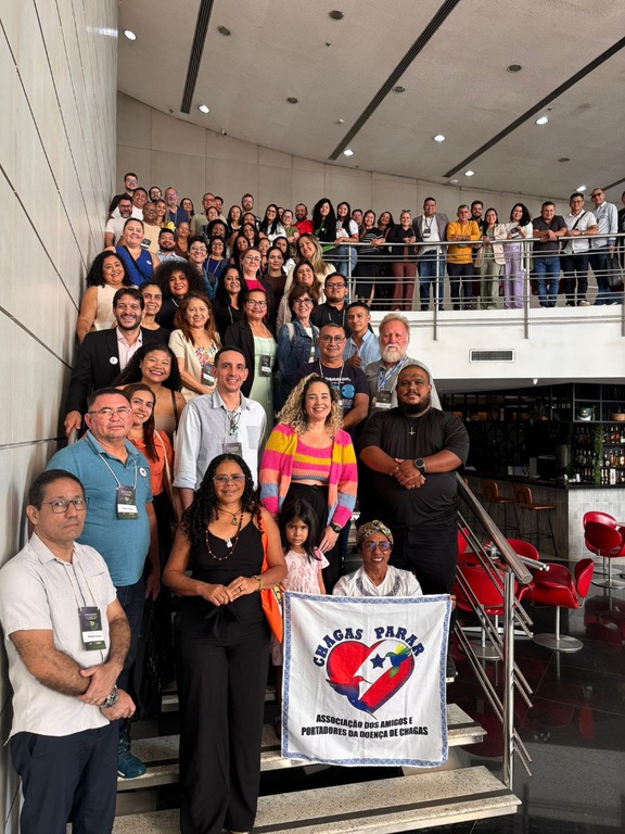 Grupo numeroso de participantes da oficina posa em uma escadaria. À frente, duas mulheres seguram bandeira da associação de Chagas.