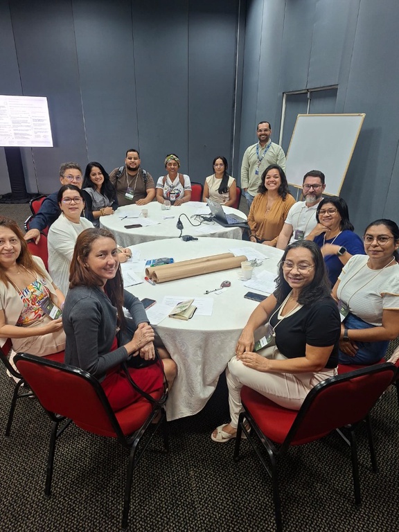 Onze pessoas sentadas ao redor de mesas redondas em sala de reunião, sorrindo para a foto durante atividade da oficina.