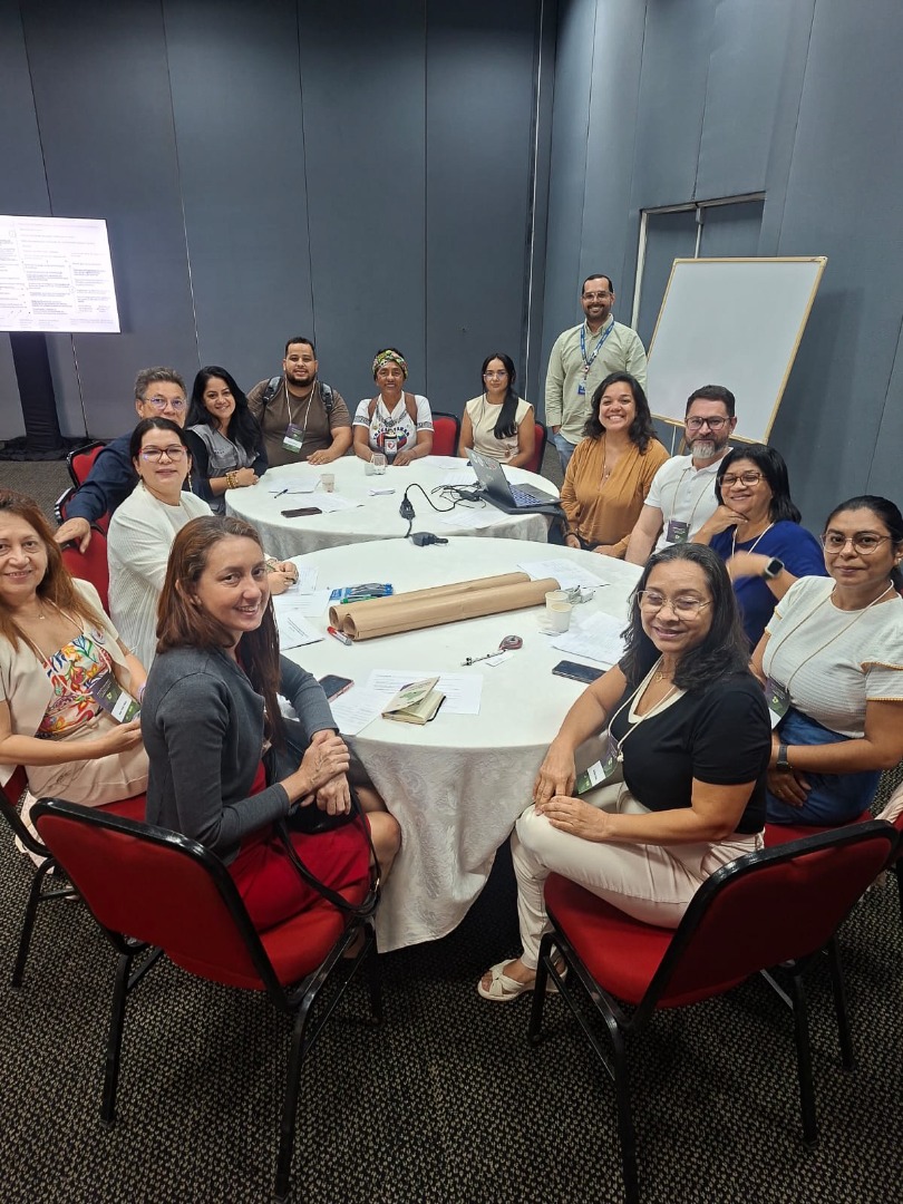 Onze pessoas sentadas ao redor de mesas redondas em sala de reunião, sorrindo para a foto durante atividade da oficina.