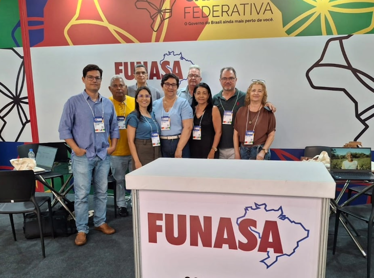 Grupo de nove pessoas posa para foto em frente a um balcão branco com o logotipo da Funasa em um estande de evento.