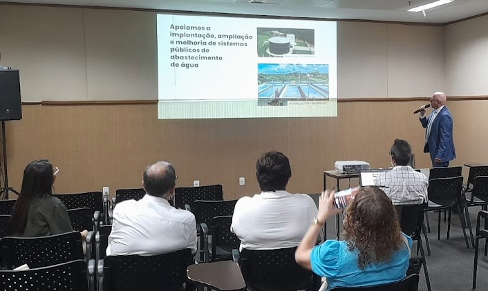Homem de terno azul apresenta slides sobre sistemas de abastecimento de água para uma pequena plateia em uma sala de conferências.