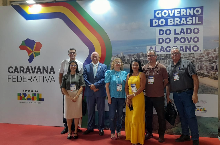 Sete pessoas posam em frente a um painel da "Caravana Federativa" com a frase "Governo do Brasil do lado do povo alagoano".