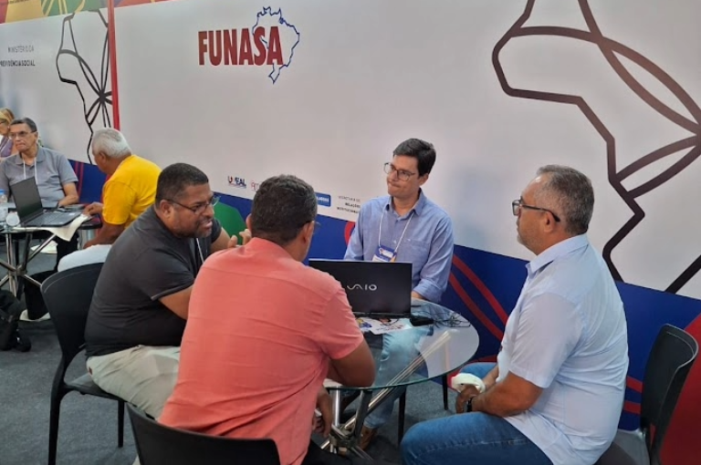 Três homens conversam sentados ao redor de uma mesa de vidro com um notebook, em um ambiente de atendimento em feira ou evento.