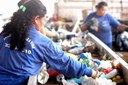 Mulher separa materiais recicláveis em esteira; ao fundo, outra trabalhadora organiza resíduos plásticos.