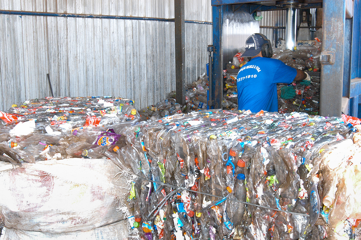 Fardos de garrafas PET prensadas; trabalhador opera prensa em galpão de reciclagem.