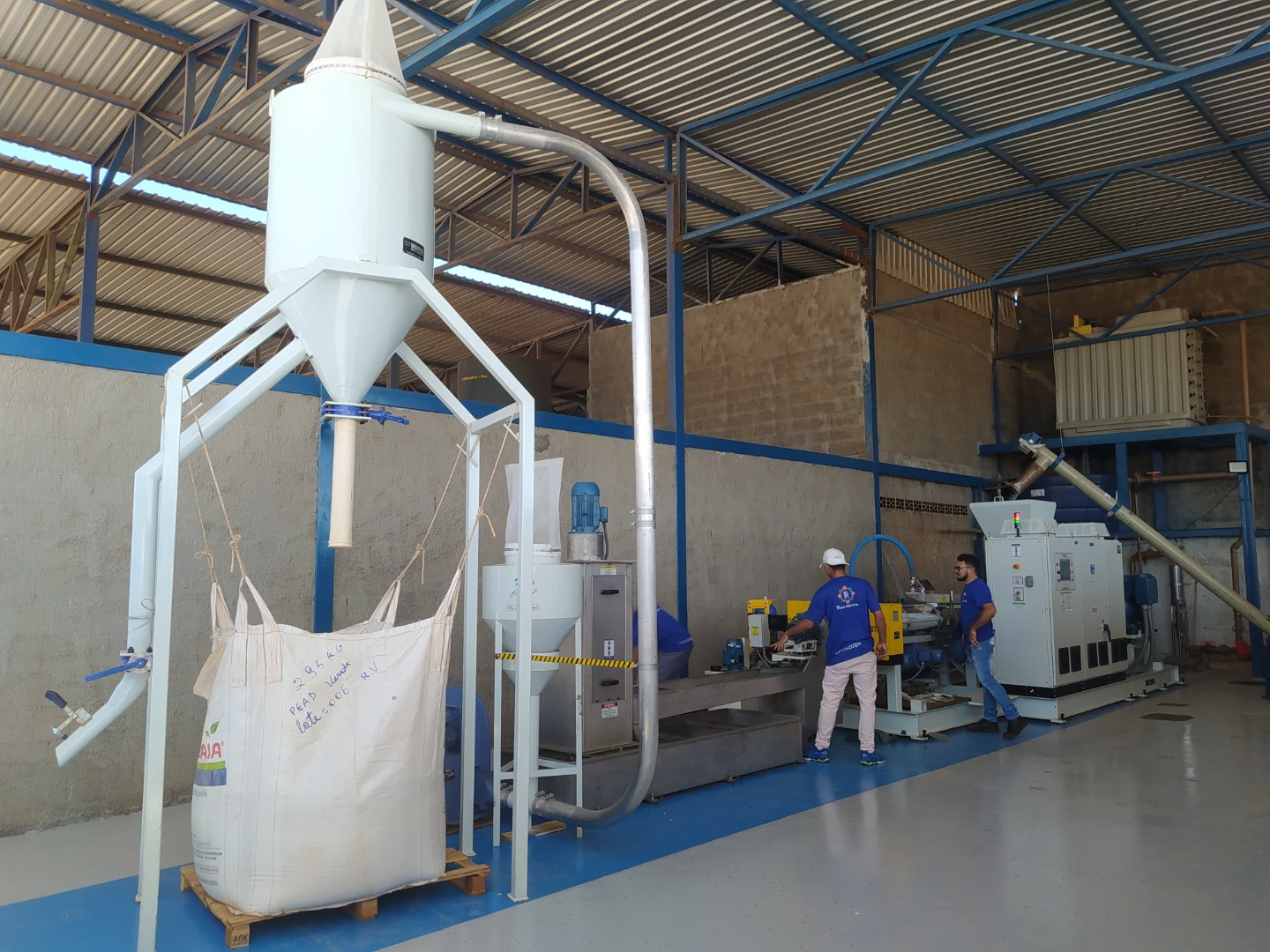 Máquina industrial de reciclagem com silo e tubulação; dois trabalhadores operam equipamentos em galpão.