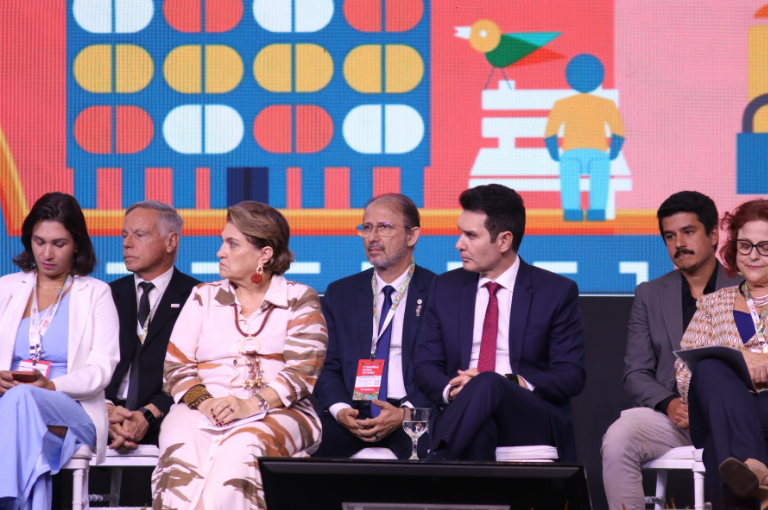 Autoridades sentadas no palco, com painel colorido ao fundo, incluindo, um homem de terno azul e óculos ao centro.