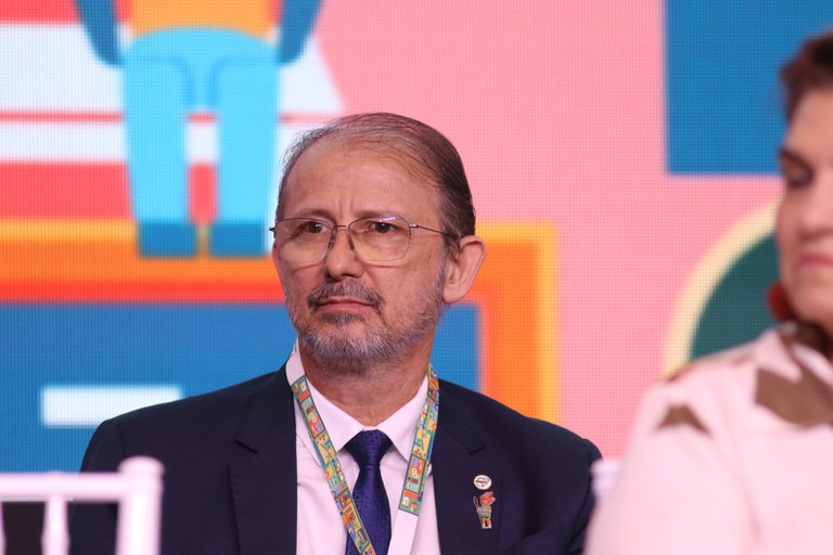 Close de Alexandre Padilha, presidente da Funasa, um homem de óculos, barba grisalha e terno azul, usando crachá e pin na lapela, sentado em evento oficial.