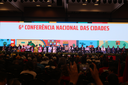 Vista ampla de auditório lotado durante a 6ª Conferência Nacional das Cidades, com diversas autoridades sentadas no palco.