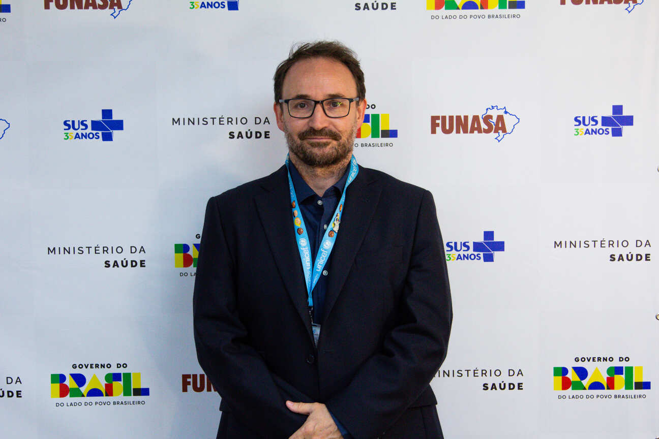 Homem branco, de óculos, terno azul escuro com crachá Unicef, com as mãos cruzada a frente, ante painel com logos da Funasa e Saúde.