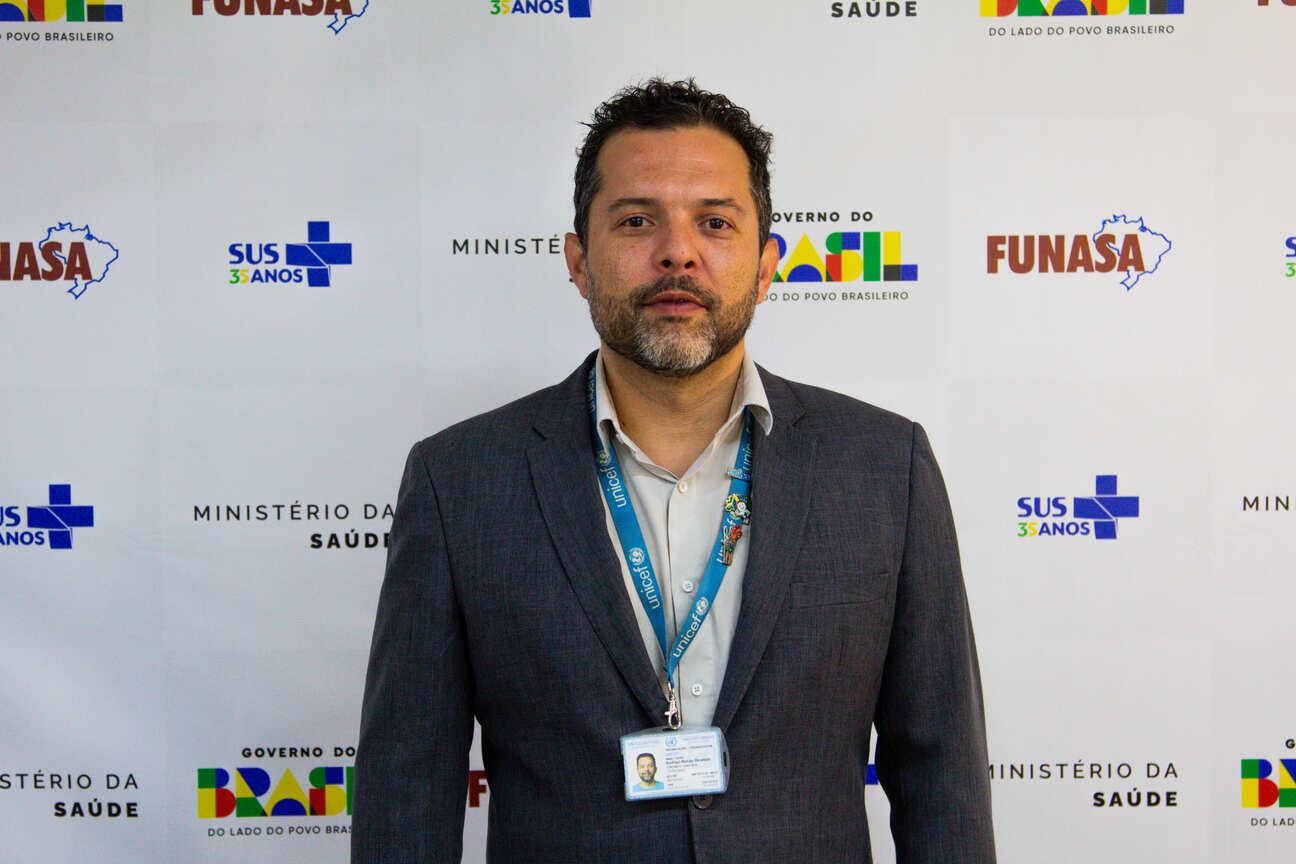 Homem pardo, de barba e terno cinza com crachá Unicef ante painel com logos da Funasa e Saúde.