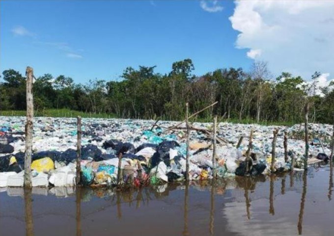 Lixão com resíduos plásticos acumulados à beira-rio, cercado por estacas de madeira, com floresta e céu azul ao fundo.