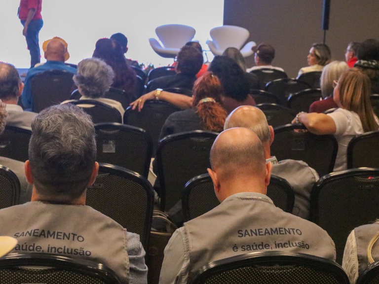 Close da parte de trás de dois coletes cinzas com a frase "SANEAMENTO é saúde, inclusão", usados por participantes em um auditório.