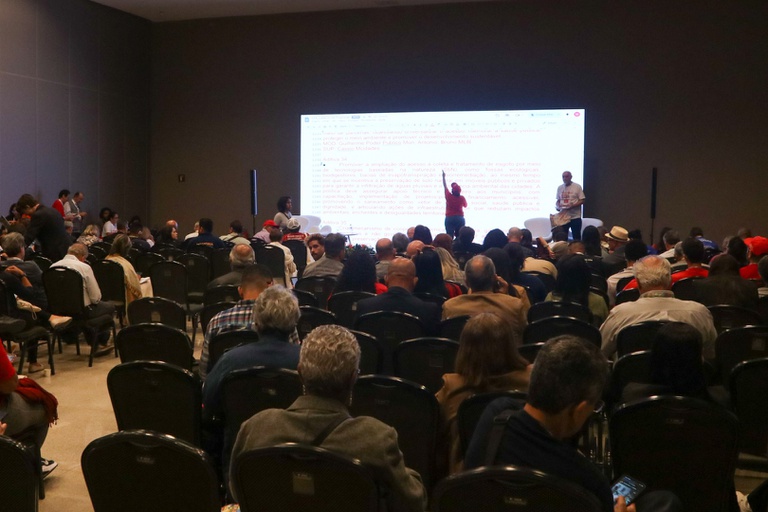 Palestrante em palco interage com o público diante de telão que exibe diretrizes sobre acesso a esgoto e tecnologias baseadas na natureza.