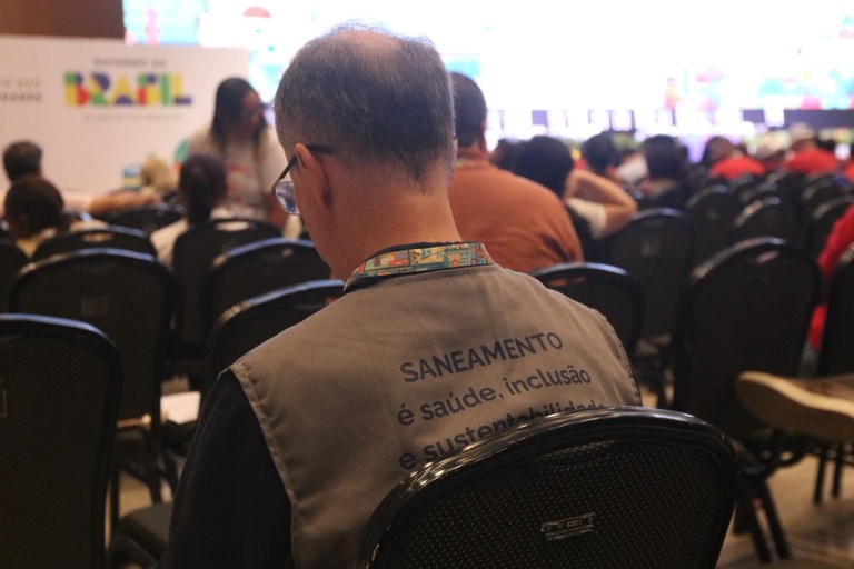 Close das costas de um homem usando colete cinza com a frase "SANEAMENTO é saúde, inclusão e sustentabilidade" em um auditório