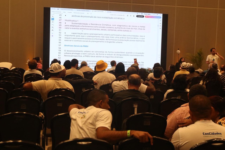 Plateia de costas observa um grande telão branco com textos sobre diretrizes de sustentabilidade e resiliência climática