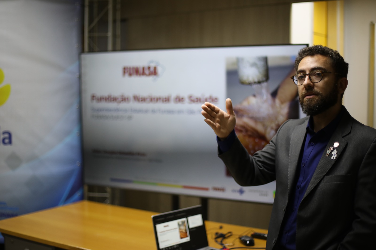 Servidor apresenta conteúdo institucional sobre saneamento básico em sala de reuniões, com apoio de tela e notebook.
