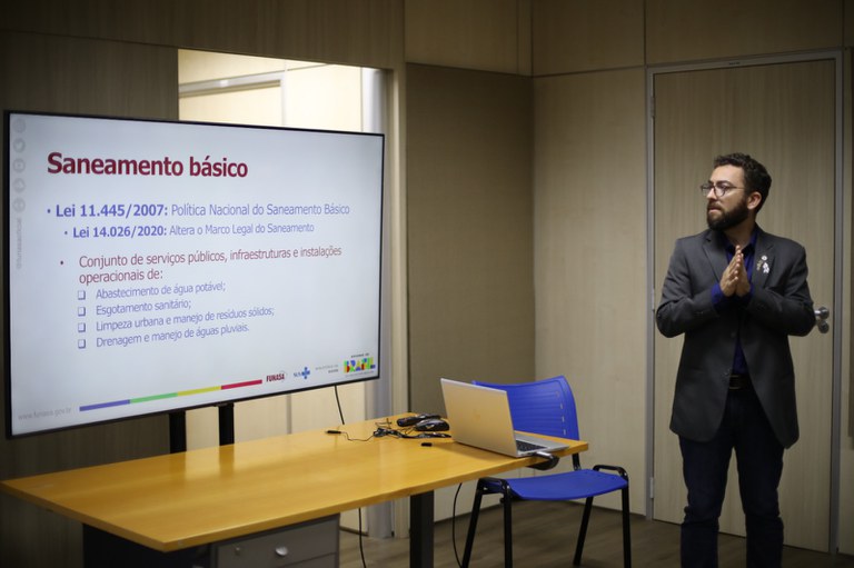 Servidor expõe conceitos de saneamento básico em apresentação institucional, com slide projetado e equipamentos de apoio.