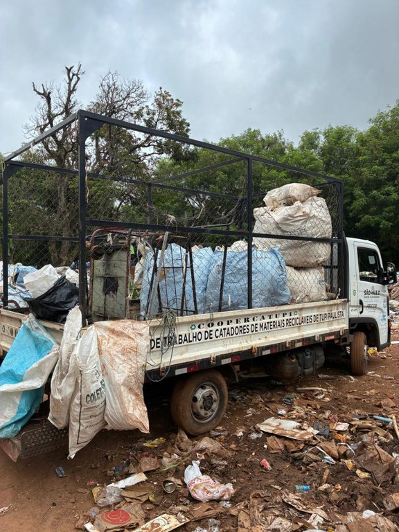 Caminhão branco antigo carregado com grandes sacos de recicláveis, em área de descarte com lixo ao redor.