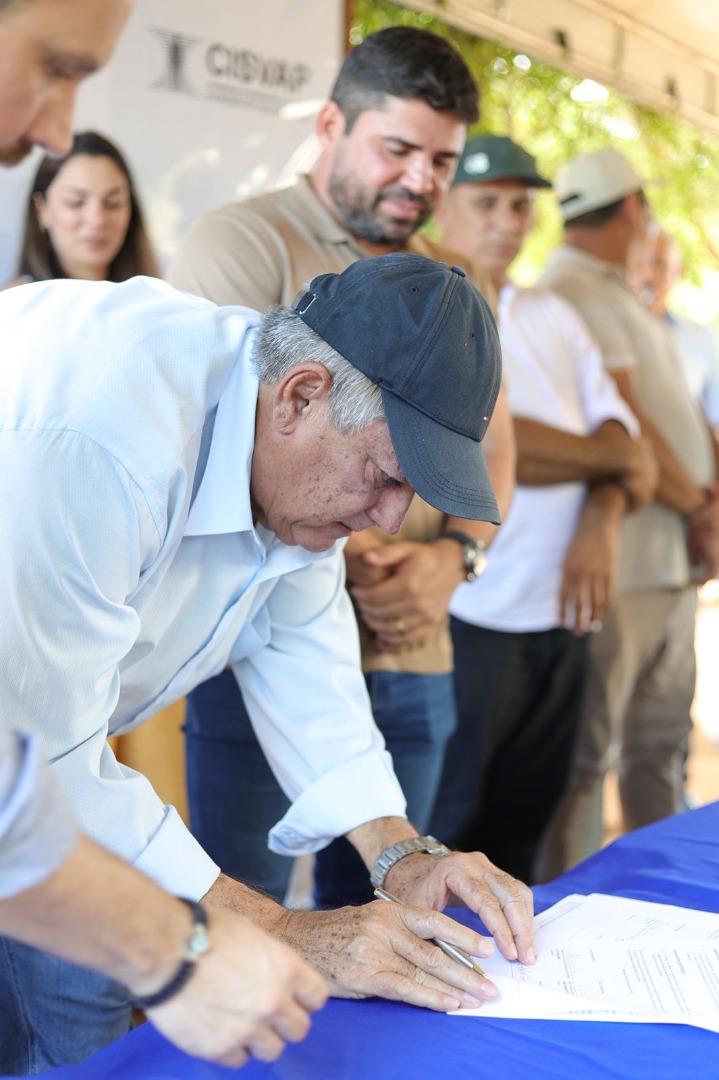 Homem idoso, de boné azul e camisa clara, assina documentos sobre uma mesa com toalha azul. - Foto_ Suest-PB_Funasa.jpeg