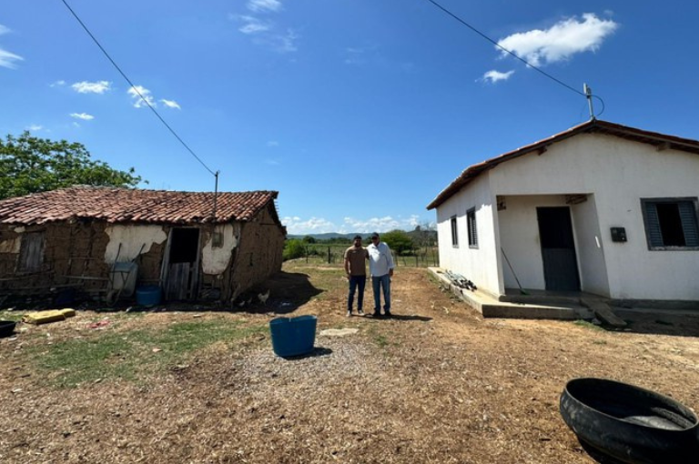 Dois homens posam entre uma casa de taipa antiga e uma casa de alvenaria nova, em terreno de terra- Foto_ Suest-PB_Funasa.jpeg
