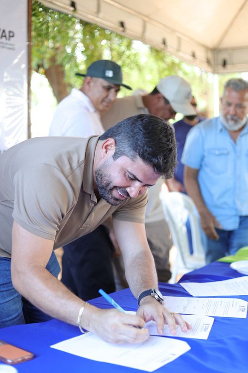 Homem jovem, de barba e camisa polo bege, assina papéis em um evento sob tenda, com outras pessoas ao fundo. - Foto_ Suest-PB_Funasa.jpeg