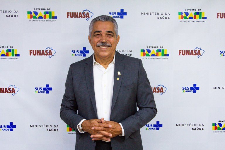 Homem branco e grisalho, de terno cinza e bigode, sorri à frente de painel com logotipos da Funasa, Ministério da Saúde e Governo do Brasil.