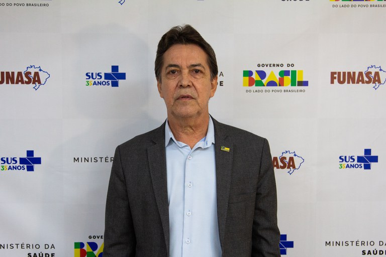 Homem de terno cinza e camisa azul clara posa diante de painel com logotipos da Funasa, Ministério da Saúde e Governo do Brasil.