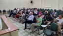 Pessoas sentadas em auditório, acompanhando reunião ou palestra, com cadeiras verdes, ar-condicionado e projetor no teto.