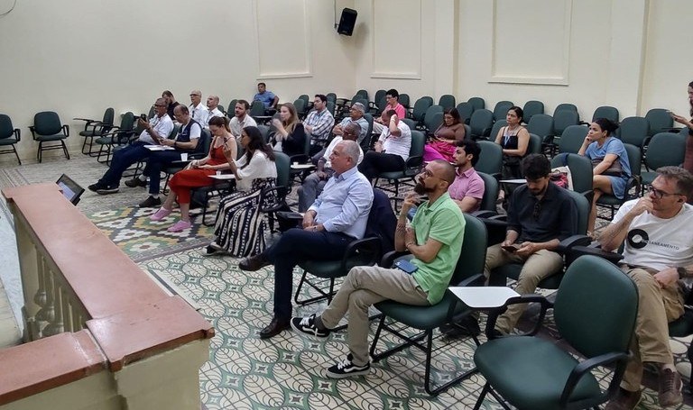 Pessoas sentadas em auditório, acompanhando reunião ou palestra, com cadeiras verdes, ar-condicionado e projetor no teto.