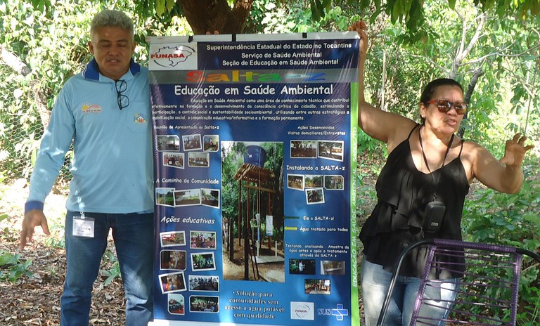 Servidores da Funasa realizam atividade de educação em saúde ambiental ao ar livre, apresentando banner institucional a moradores da comunidade.