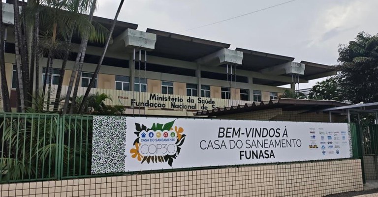Fachada da Funasa com faixa “Bem-vindos à Casa do Saneamento – COP30”, palmeiras ao redor e prédio institucional ao fundo.