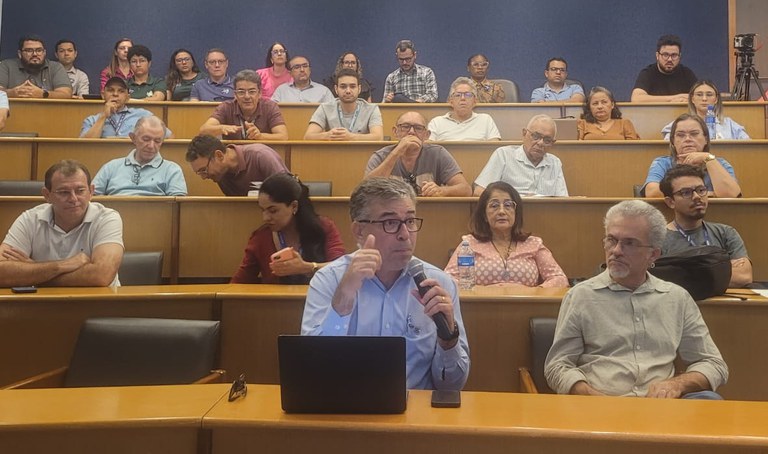 Auditório cheio com participantes sentados em níveis; homem à frente fala ao microfone com notebook aberto, enquanto o público acompanha.