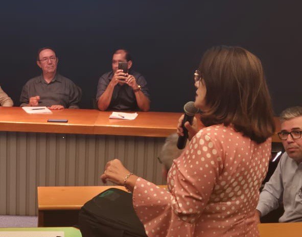 Mulher de pé fala ao microfone em auditório, voltada à mesa de autoridades, durante reunião ou debate institucional.