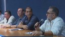 Mesa de reunião com homens sentados; um deles fala ao microfone enquanto os demais acompanham a discussão em ambiente institucional.
