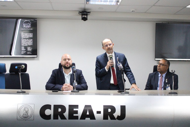 Alexandre Motta fala ao microfone em mesa do CREA-RJ, ao lado do presidente Miguel Fernández e do superintendente da Funasa, Robson Maia.